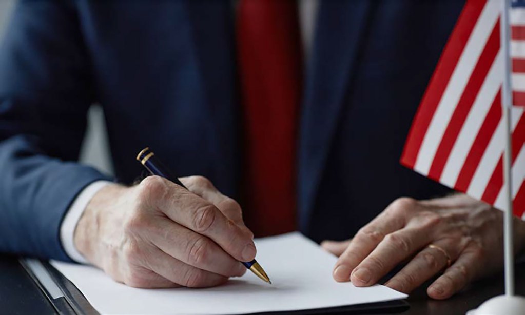 Un hombre vestido con traje y corbata firmando un documento, con una bandera estadounidense visible detrás de él.